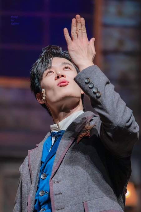A performer with dark hair wearing a gray suit with a blue vest and bow tie, looking upward with one hand up, tagged 'Pungi to Eighteen'.