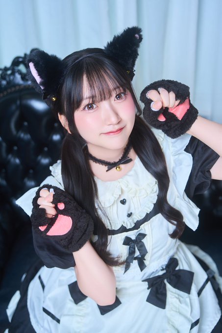 A young woman poses wearing a black and white maid-style outfit with black cat ears and matching paw gloves, seated against a black tufted leather background.