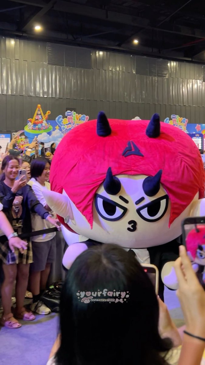 A large pink and white mascot character with horns and an angry expression surrounded by a crowd taking photos at what appears to be a convention or event, with a watermark reading 'yourfairy'.