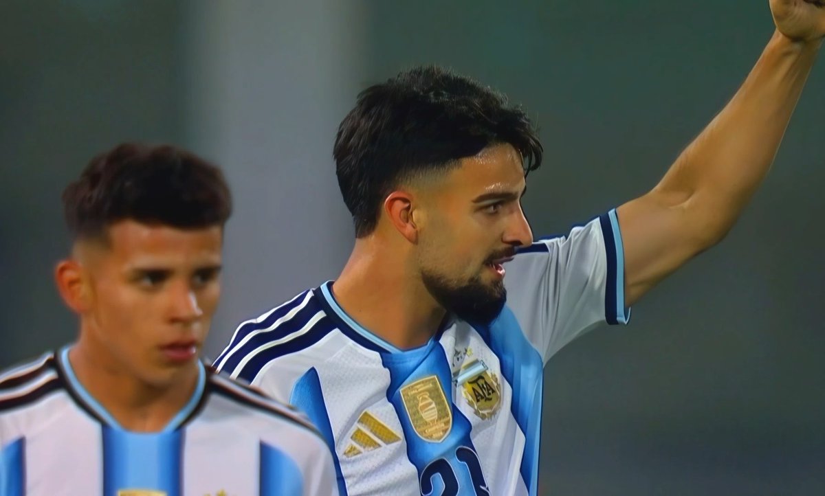 An Argentine national soccer player with a beard and jersey number 21 raises his arm in celebration, while a teammate in a light blue and white striped jersey stands in the foreground.