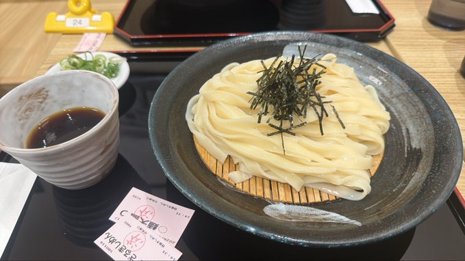 A serving of cold udon noodles topped with shredded nori seaweed on a wooden plate, accompanied by a small bowl of dipping sauce and garnishes, served on a black tray with a paper slip.