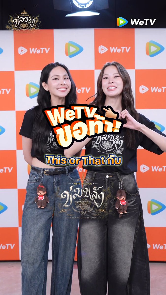 Two women wearing matching black t-shirts and jeans smile brightly while posing together at a WeTV press event, with promotional graphics reading "WeTV ขอท้า!" and "This or That กับ" overlaid on the image for the Thai series "ข่มขลัง" (Khom Khlang The Series).