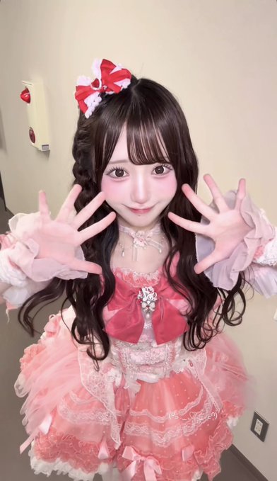 A young woman in a pink frilly dress with red bows and a matching hair bow poses with her hands raised.