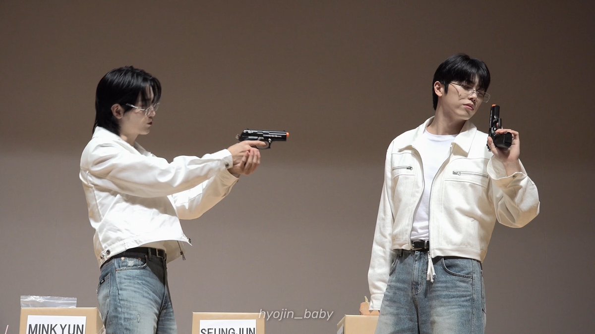 Two men pose on stage in matching white jackets and jeans, one aiming a prop gun while the other holds one near his face, with name placards reading "MINKYUN" and "SEUNG JUN" visible at the bottom—capturing the playful "noir scene" concept from the Tokyo release event for OnNOff.