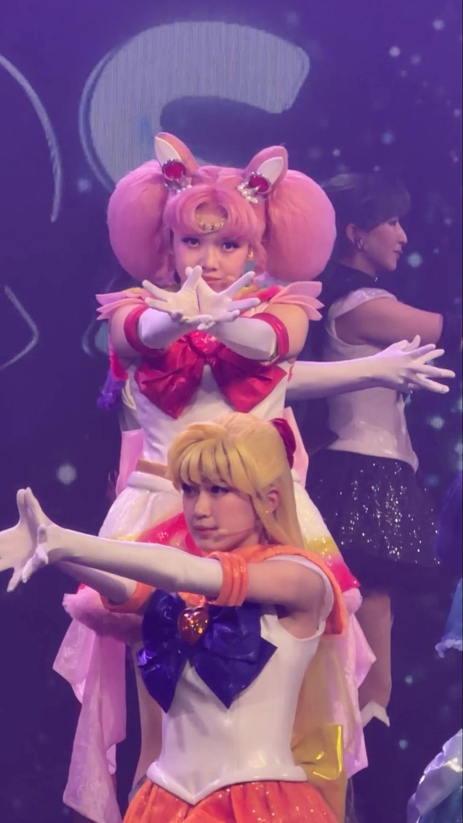 Two performers in Sailor Moon cosplay striking a crossed-arm pose on stage, with the foreground performer dressed as Sailor Venus (orange) and the performer behind her as Sailor Chibi Moon (pink).