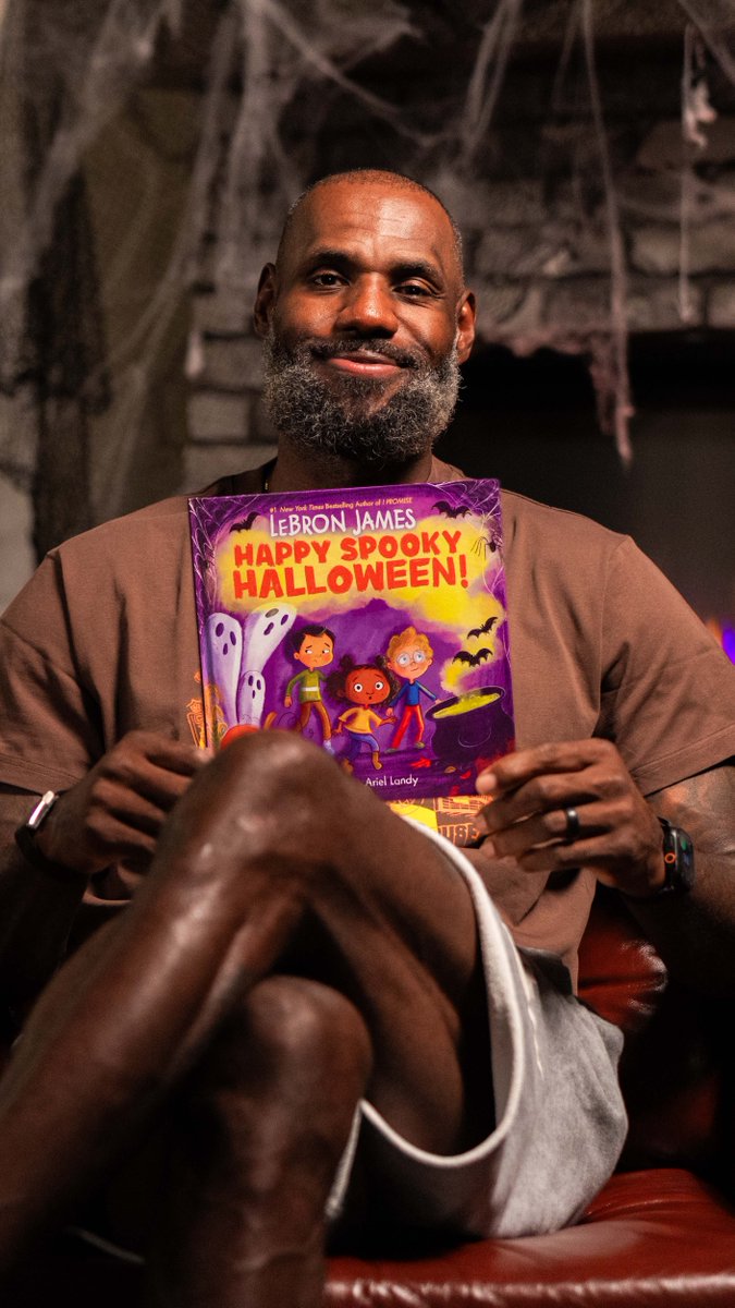 LeBron James sits smiling on a leather couch in a Halloween-themed setting with cobwebs, holding up his children's book titled "Happy Spooky Halloween," featuring colorful illustrations of kids in costumes on the cover.