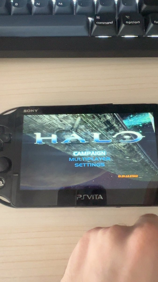A handheld Sony PS Vita displays the main menu of a "Halo" game port featuring Campaign, Multiplayer, and Settings options, positioned below on a desk beneath a black keyboard. The image illustrates the work-in-progress recompilation project referenced in the caption, showing the iconic Halo title screen successfully running on Sony's handheld console.