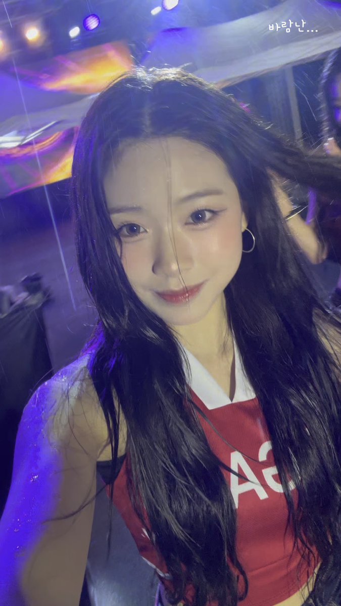 A young woman with long dark hair and glamorous makeup smiles for a selfie in a venue with blue and purple stage lighting, wearing a red and white crop top with partial lettering visible; Korean text '바람난날' (meaning 'windy day') appears in the upper right corner.