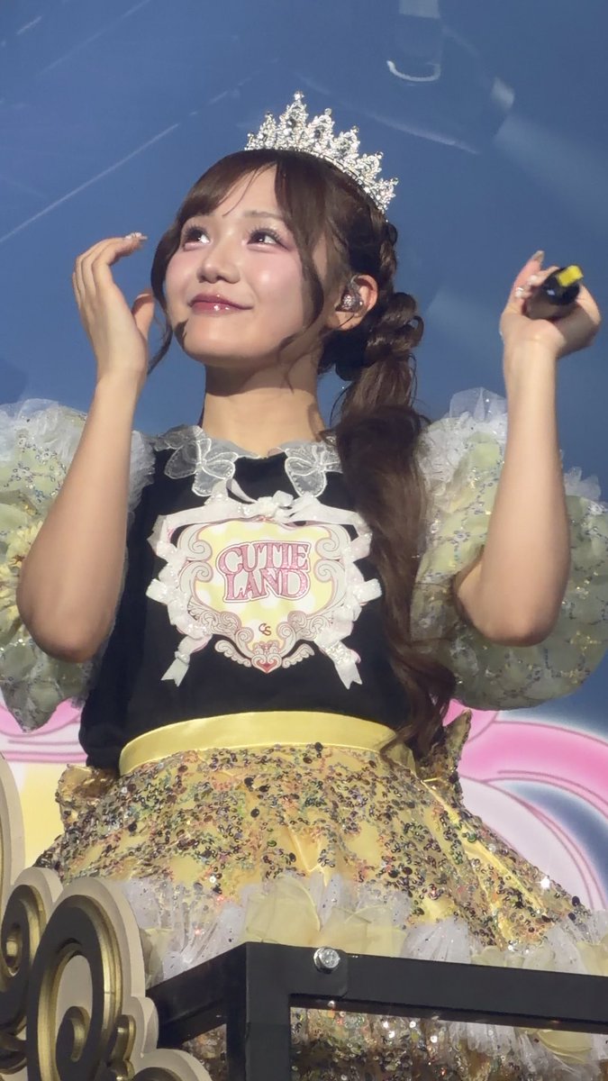A performer wearing a silver tiara and a 'Cutie Land' themed costume with a yellow ruffled skirt looks upward on stage, smiling.