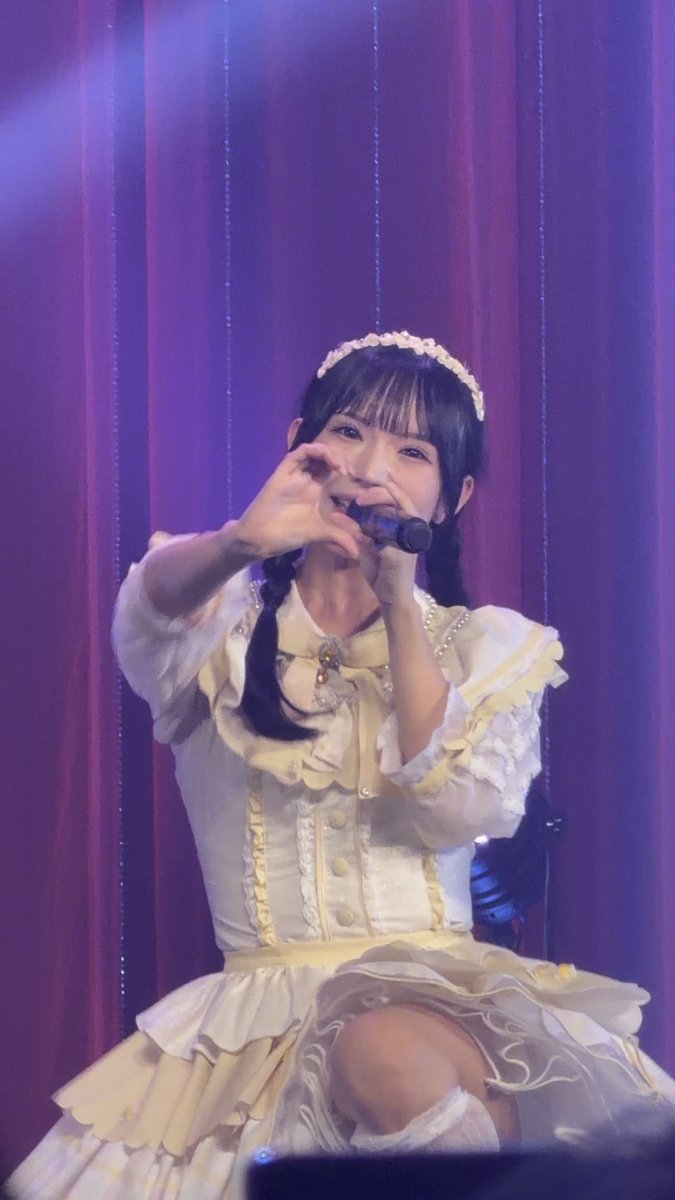 A young woman wearing a white and yellow dress with a small crown and pigtails performs on stage with a purple curtain backdrop, making a heart shape with her hands while holding a microphone.