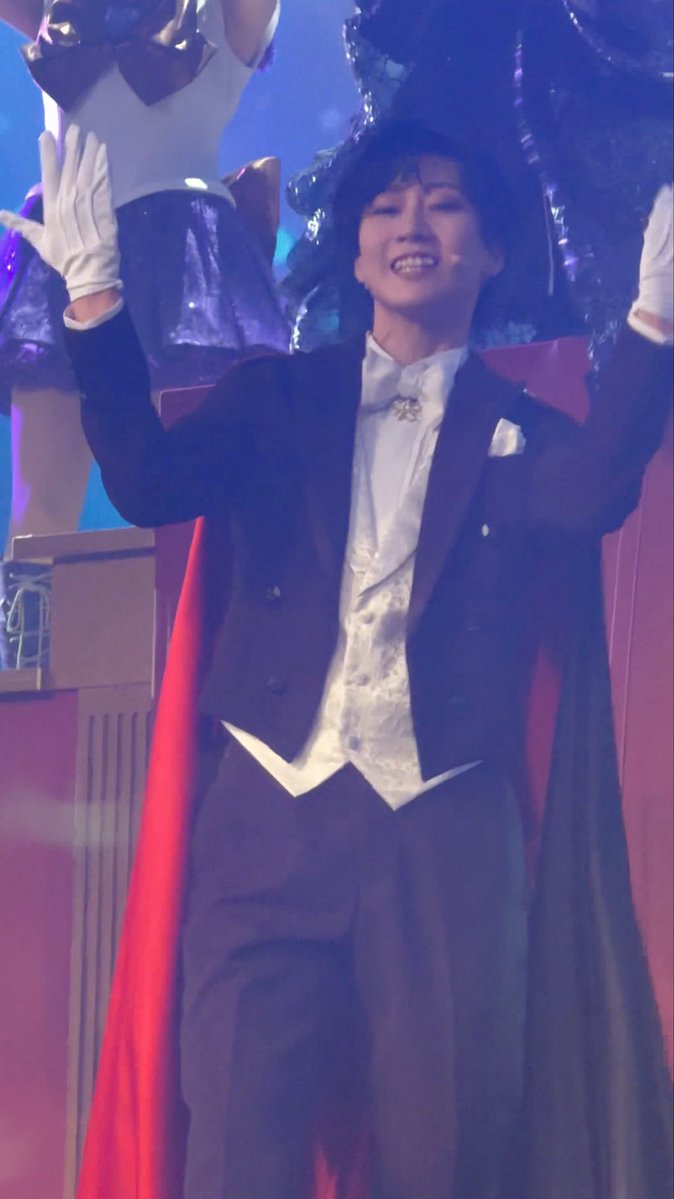 A young man in a black tuxedo with a white vest, cape, and gloves, smiling broadly with his arms raised on a theatrical stage set.