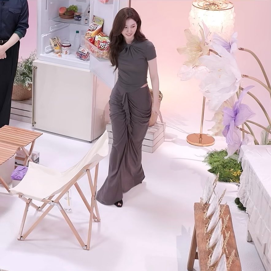 A woman with long brown hair wearing a sleek gray top and a draped gray skirt stands in a pink and white set featuring a refrigerator with snacks, a chair, and decorative flowers.