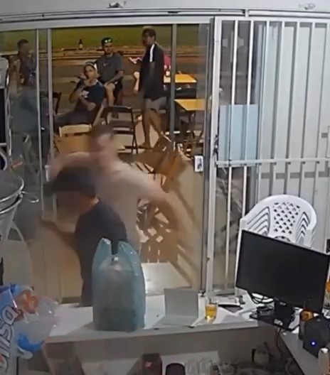 The image, likely a security camera screenshot, shows a chaotic scene through a glass window where a blurred figure appears to be attacking others in what looks like a restaurant or cafeteria setting, with several people visible in the background. In the foreground, there's a desk with a monitor and a large water jug. The violent, sudden attack depicted relates to the post's joke about the "cuck" character eliminating everyone the piranha endorses, evoking the relentless assassin storyline of John Wick.