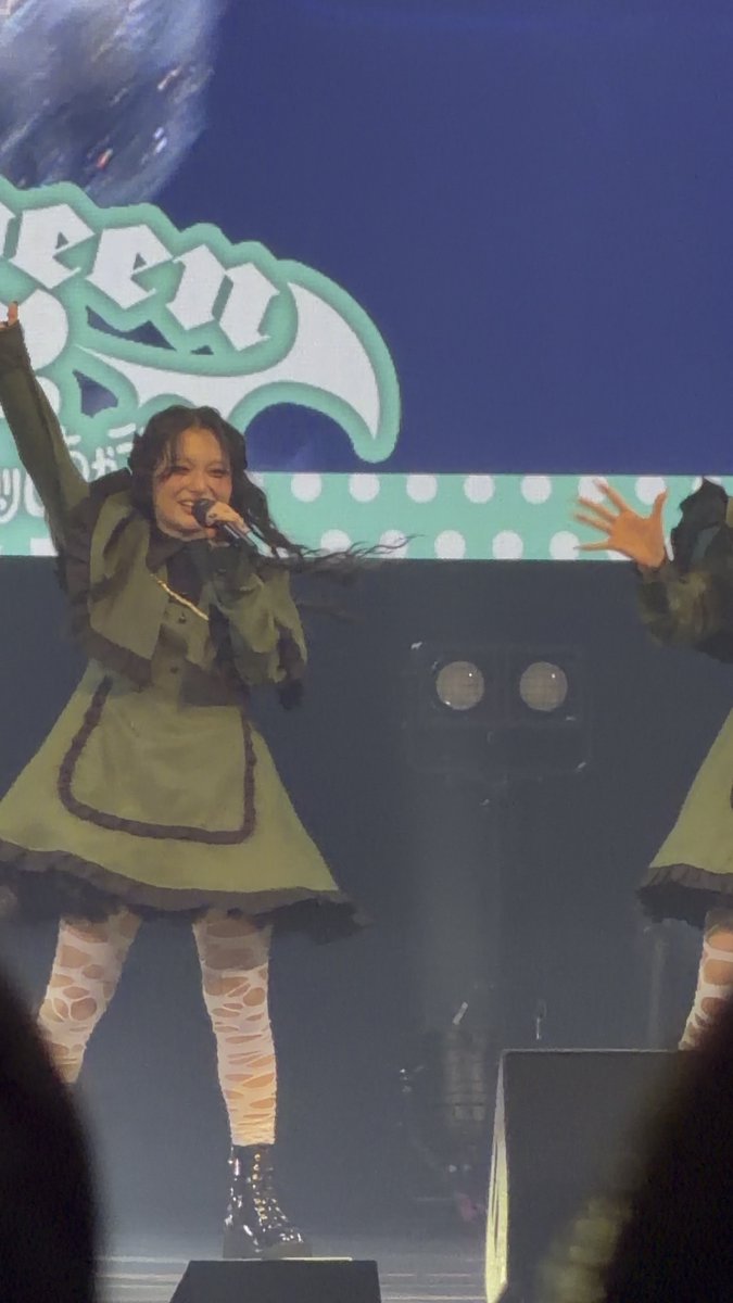 A performer in a green ruffled dress with patterned tights sings into a microphone on stage with a green graphic logo backdrop.