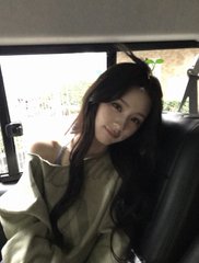 A young woman with long dark hair wearing a beige jacket sits inside a vehicle, smiling at the camera.
