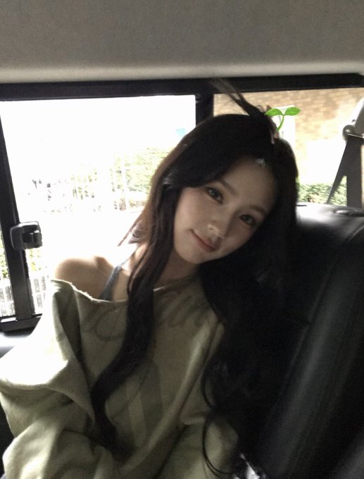A young woman with long dark hair wearing an olive off-shoulder top sitting inside a vehicle.