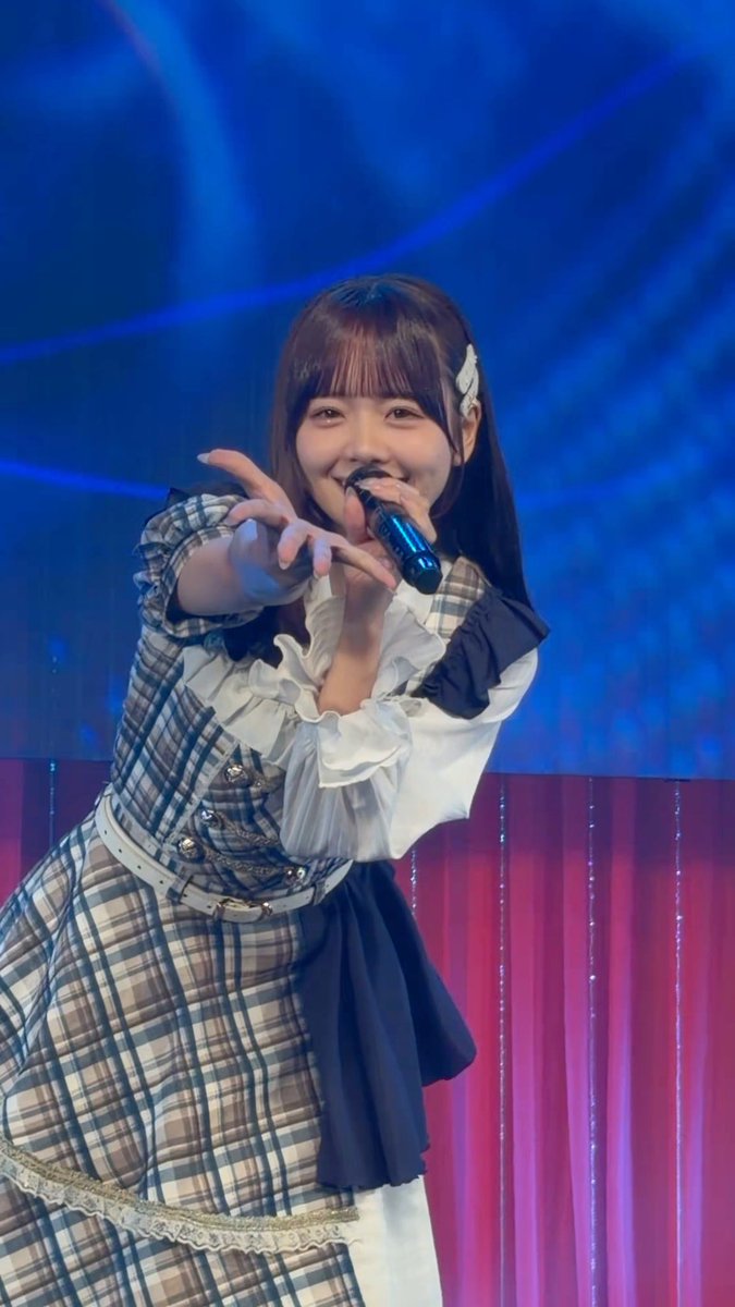 A young woman performs on stage in a plaid dress with white ruffled sleeves, pointing toward the camera while holding a microphone against a blue-lit backdrop.