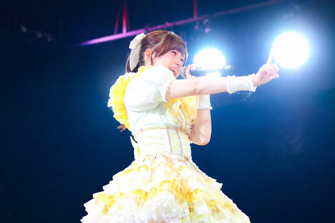A young woman in an elaborate yellow and white frilly dress performing on a dimly lit stage with bright spotlights behind her.