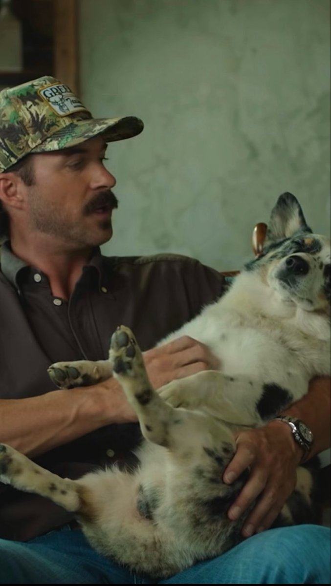 A man with a mustache wearing a camouflage cap and brown shirt, holding a spotted dog upside down on his lap as if giving it a belly rub.