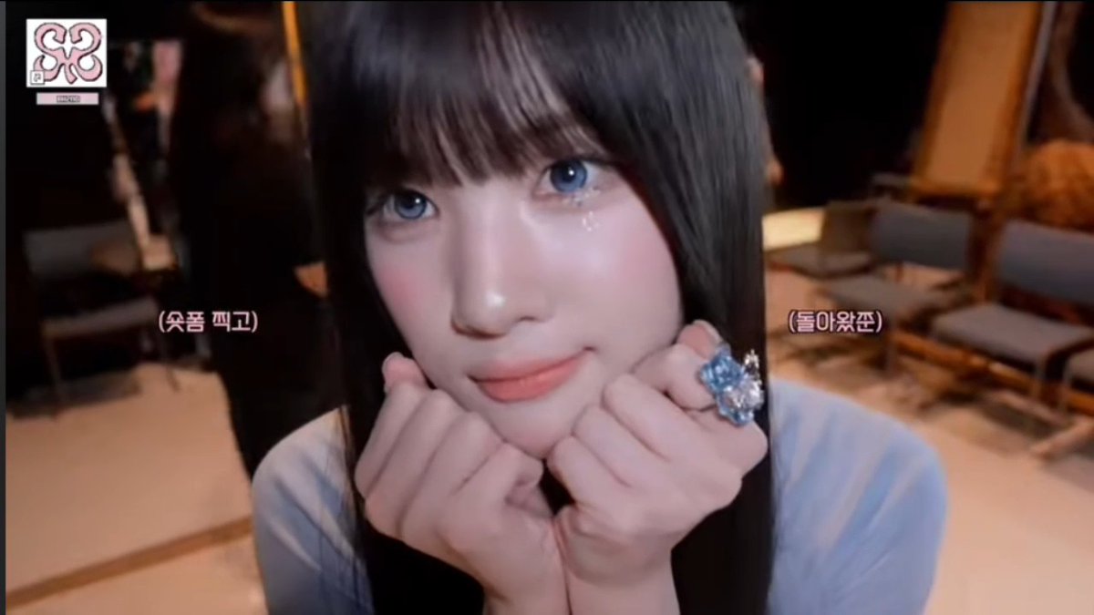A close-up of a smiling woman with dark hair and bangs, framed by her hands near her face, showing a large blue ring. Korean subtitles read '(웃음 뼈고)' and '(돌아왔준)' with a Chanel logo in the top left corner.