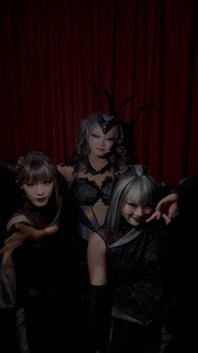 Three costumed performers from the Shining Theater production pose dramatically in elaborate dark, gothic-style outfits with spiked headpieces and lace details, set against a deep red curtain backdrop.