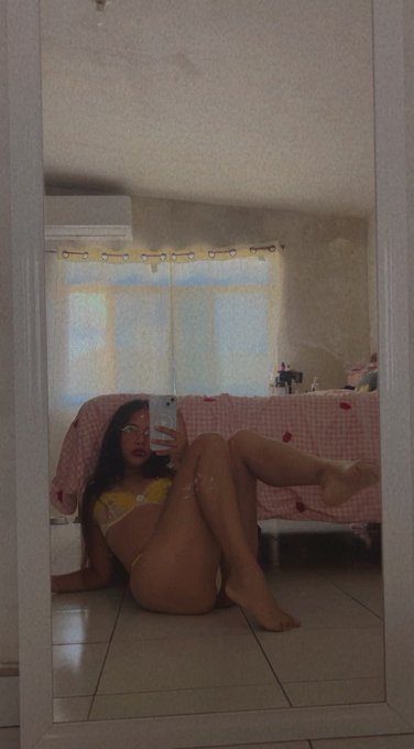 A mirror selfie of a young woman in a yellow bikini sitting on a tiled floor, holding a phone to capture the reflection.