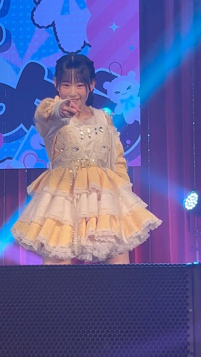 A young woman wearing a yellow and white frilly dress and decorative headpiece points at the camera with a smile while standing on a stage with colorful lighting.