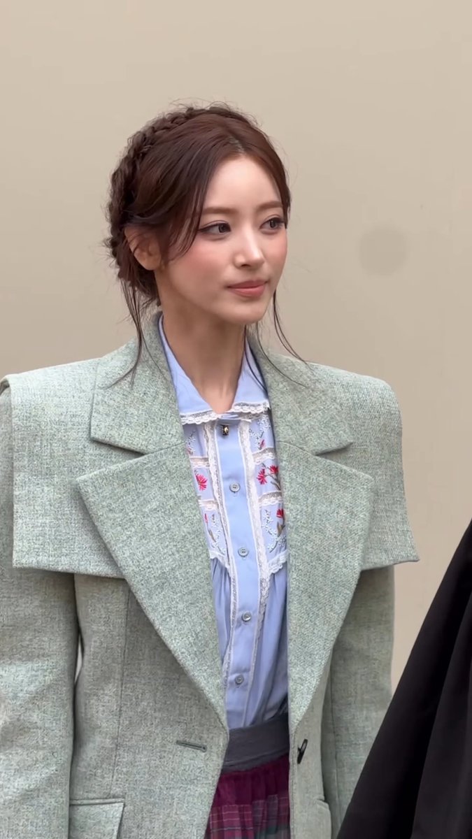 A young woman, referred to as "Princess Chloe," is depicted wearing a stylish sage green wool coat layered over a light blue blouse adorned with delicate floral embroidery and lace trim, paired with a plaid skirt. Her hair is styled in an elegant updo with loose tendrils framing her face, complementing her soft, graceful expression.