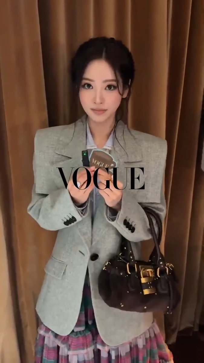 A woman in a gray blazer and pleated skirt holds a 'VOGUE' branded item, with a black handbag on her shoulder, standing before a curtain backdrop with 'VOGUE' text overlay.