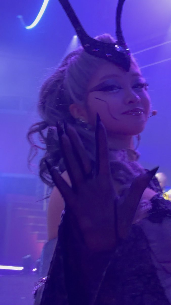 A performer on stage wears a dark horned headpiece and theatrical makeup with sharp, pointed fingernails visible on a raised hand, all bathed in purple stage lighting.