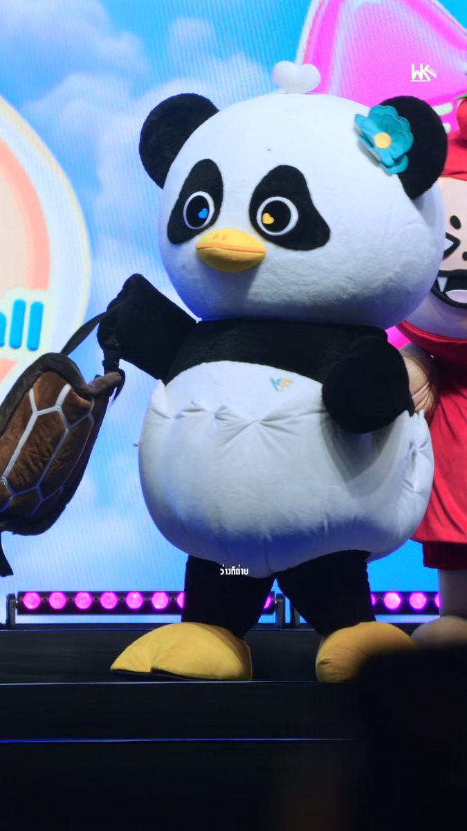 A large mascot costume of a black and white panda wearing a blue flower accessory, standing on a stage with pink lights.