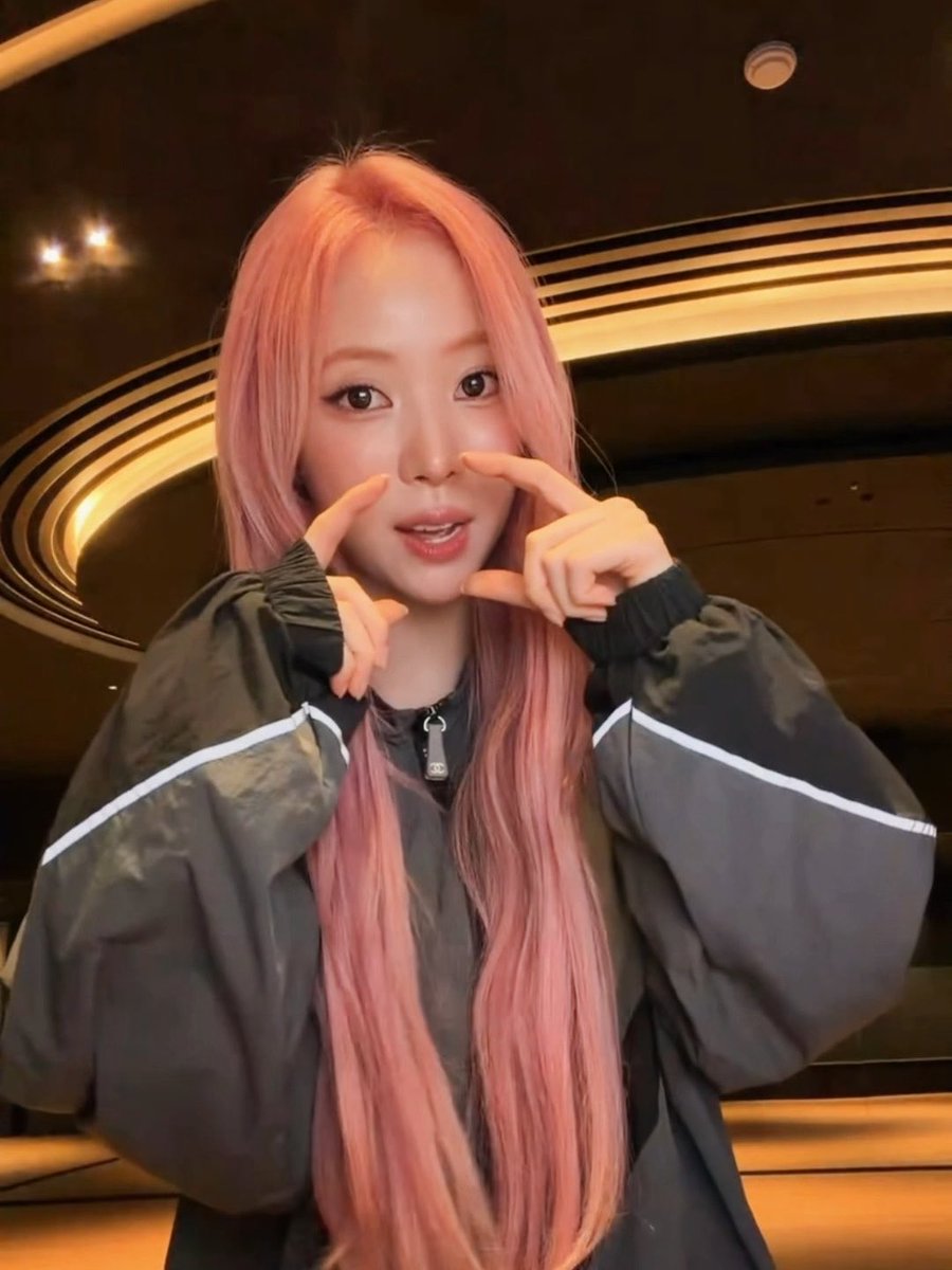 A young woman with long pink hair wearing a black jacket with white piping, making a finger frame gesture around her face in what appears to be an upscale venue with ornate ceiling lighting.