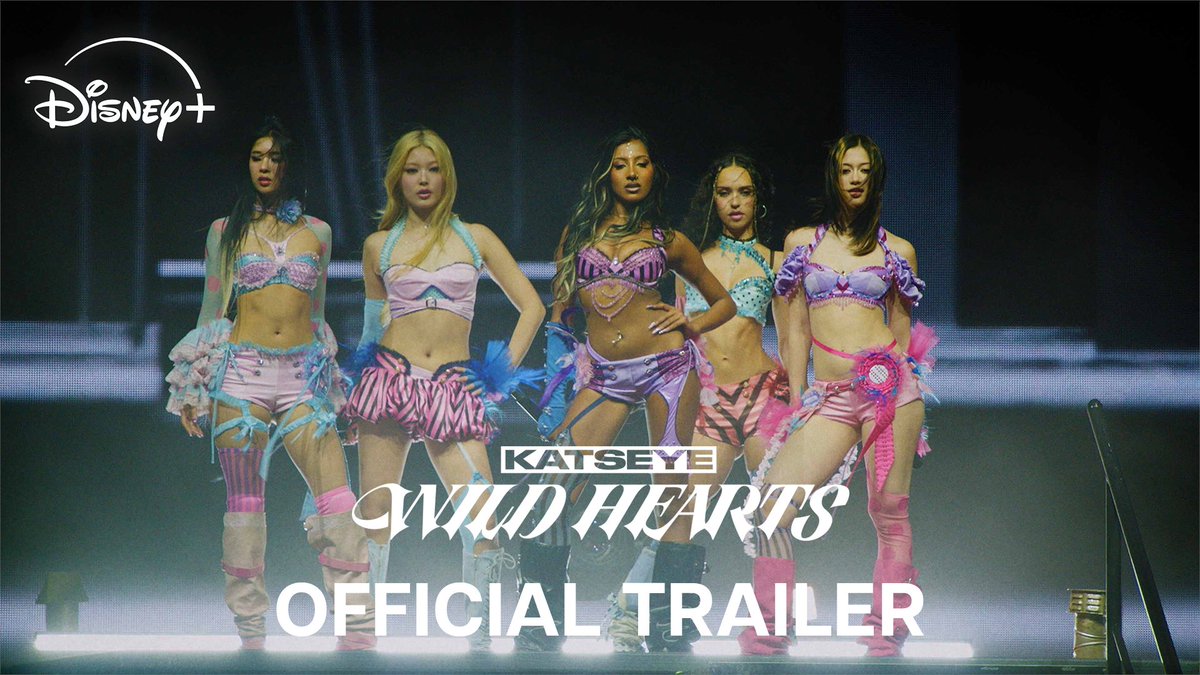 Five members of KATSEYE pose in coordinated pastel pink and blue stage outfits on a dark stage, with text overlays promoting the "KATSEYE: WILD HEARTS OFFICIAL TRAILER" and the Disney+ logo, visually matching the post's announcement of the girl group's documentary film on Disney+.
