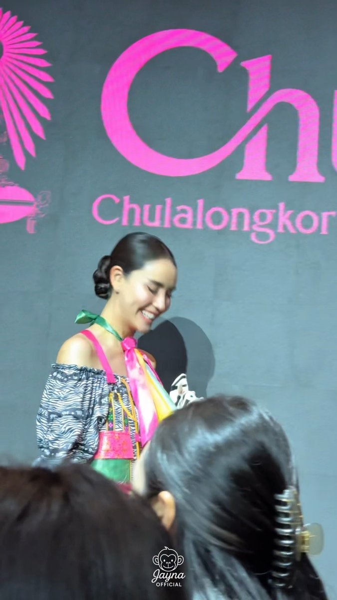 A young woman with her hair pulled back smiling on stage in front of a large pink backdrop with the text 'Chulalongkorn' partially visible.