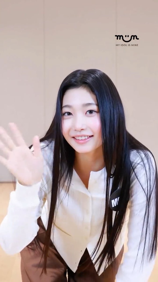 A young woman with long black hair smiles and waves at the camera, wearing a white shirt and brown pants, with a 'MY IDOL IS MINE' (MIIM) logo in the corner.