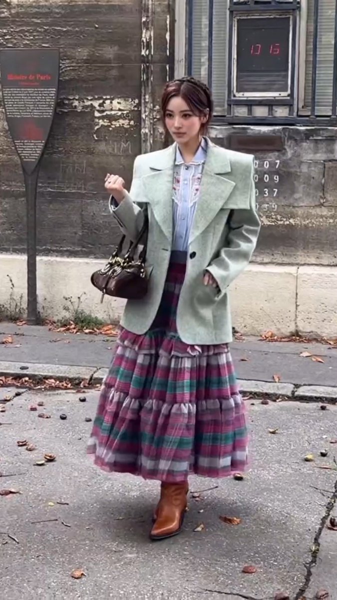 A young woman poses on a Paris street in a mint green blazer, a light blue ruffled shirt, a long plaid tiered skirt in pink, green, and white, brown boots, and carrying a dark brown handbag.