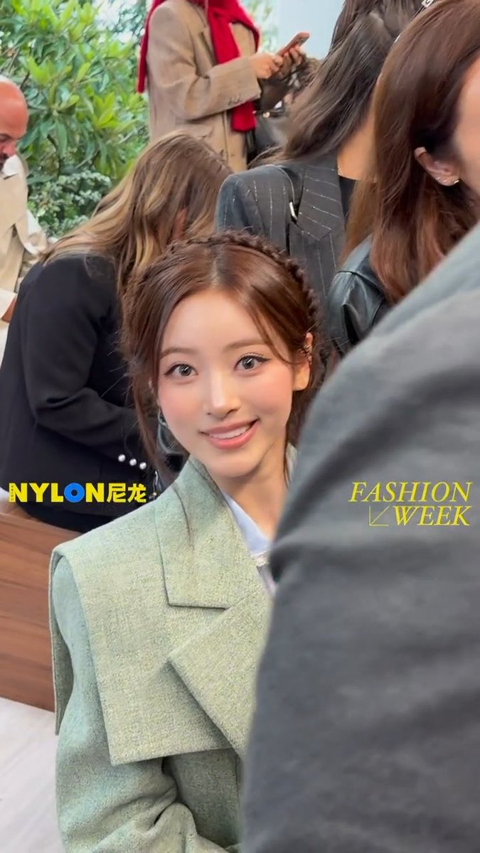 A young woman with braided hair wearing a green coat smiles at the camera at what appears to be a fashion event, marked with "NYLON 尼龙" and "FASHION WEEK" watermarks.
