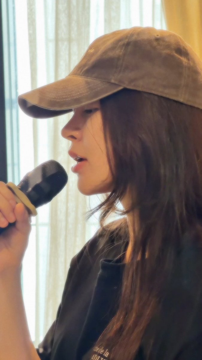 Profile close-up of a woman wearing a brown corduroy cap speaking into a handheld microphone against a window with sheer curtains.