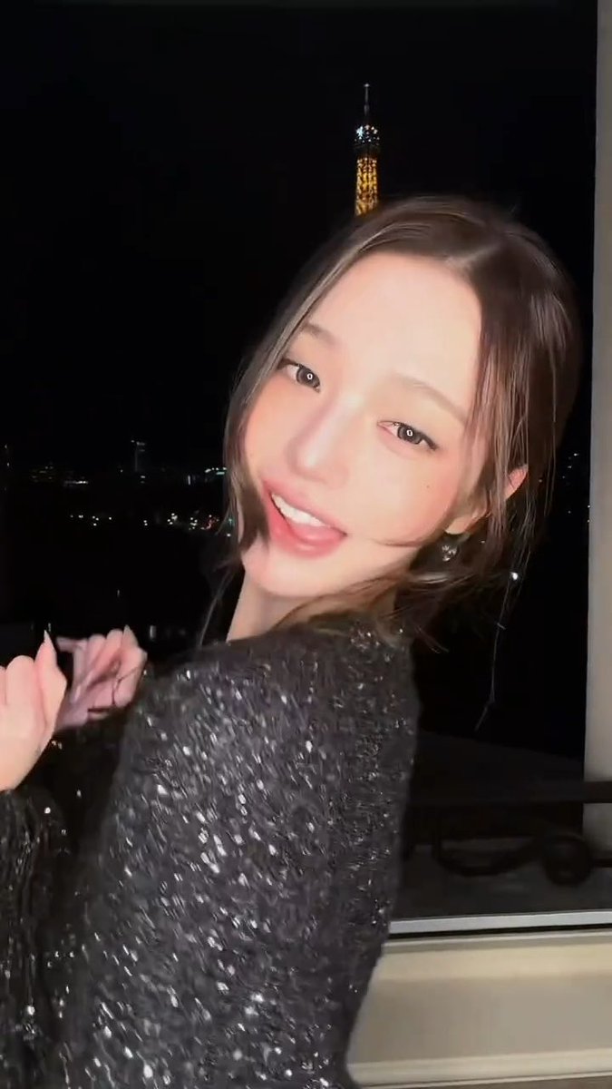 A young woman with shoulder-length hair in a sparkly black top smiling in front of a window at night, with an orange illuminated tower visible in the background.