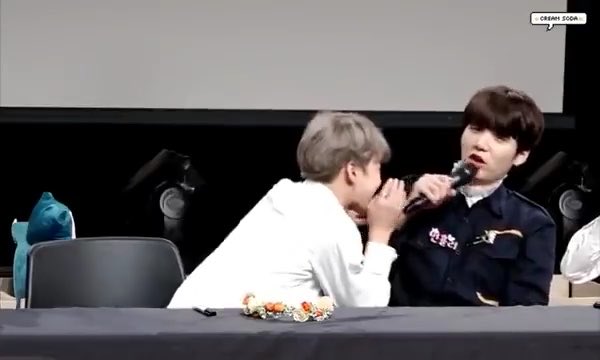 Two young men, appearing to be K-pop idols, are seated at a table; the one in white leans toward the other while the man in a dark uniform speaks into a microphone, with "CREAM 2004" visible in the upper right corner.