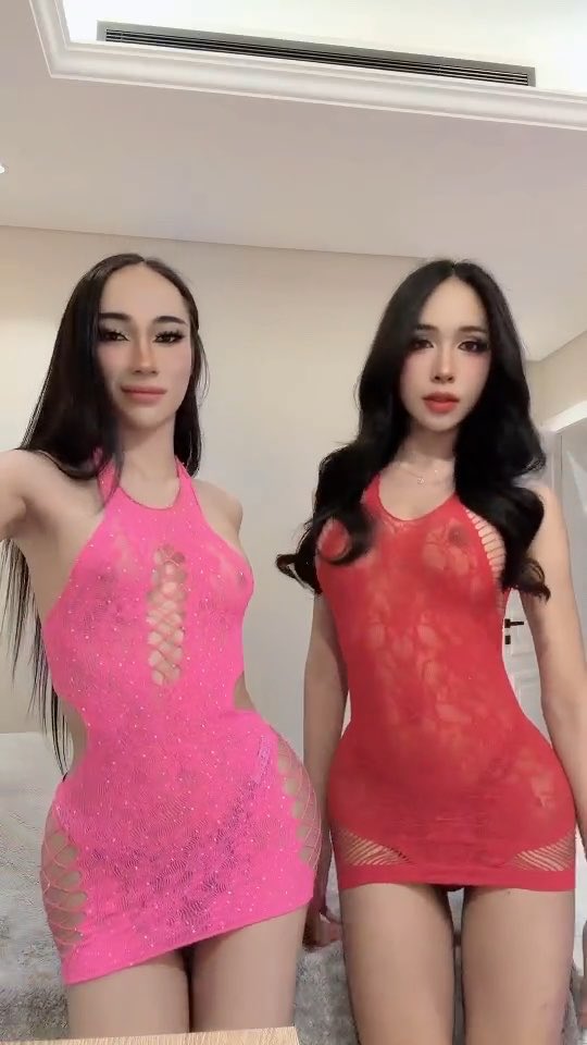 Two women pose together indoors; the woman on the left wears a pink cutout mini dress, while the woman on the right wears a red sheer lace dress.