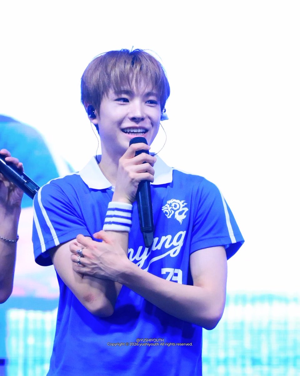 A young male K-pop performer with blonde hair smiles while holding a microphone, wearing a blue and white sports jersey, with the watermark '@YUSHIYOUTH' at the bottom.