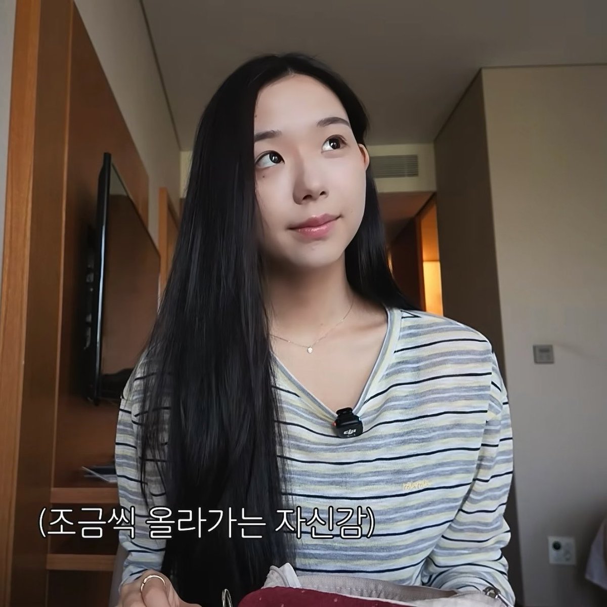 A young woman with long black hair wearing a striped v-neck shirt in a hotel room, with Korean subtitle '(조금씩 올라가는 자신감)' meaning 'confidence that rises little by little'.