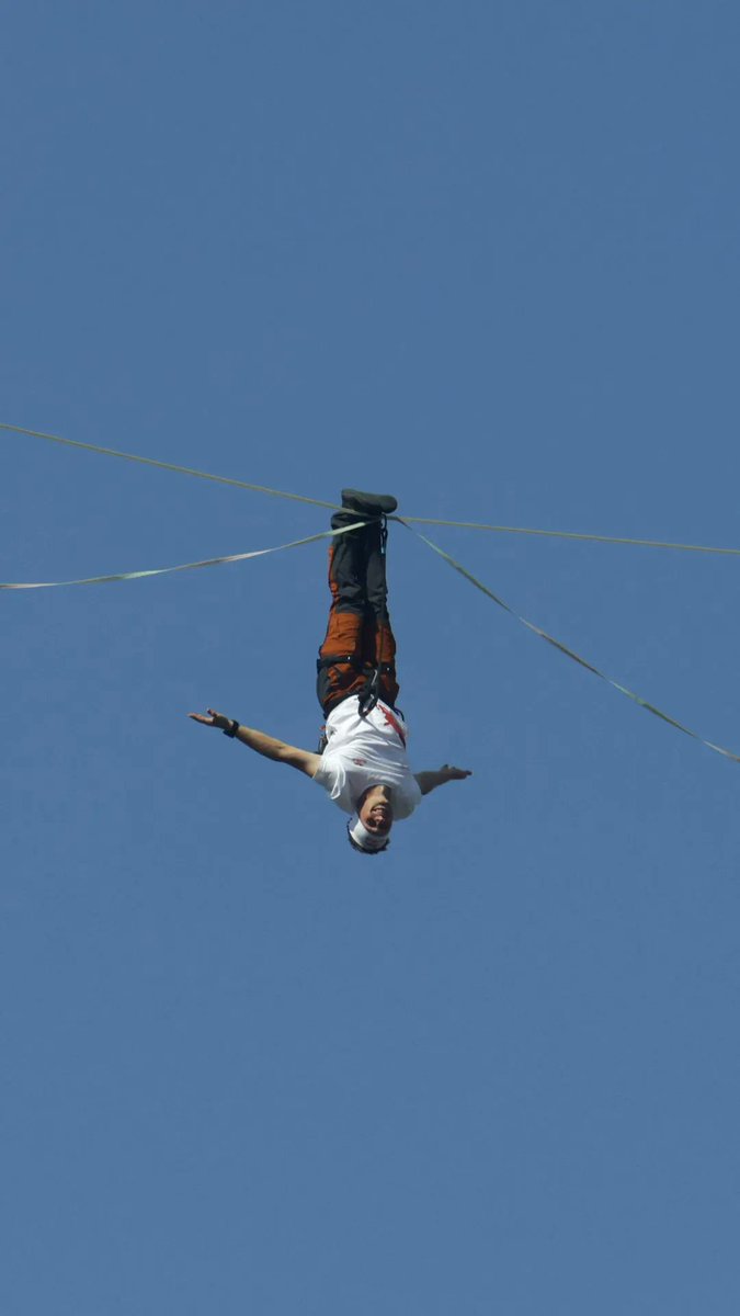A slackliner balances upside down on a thin line high against a clear blue sky, performing an acrobatic move during a daring high-wire walk between two skyscrapers in central Seoul.
