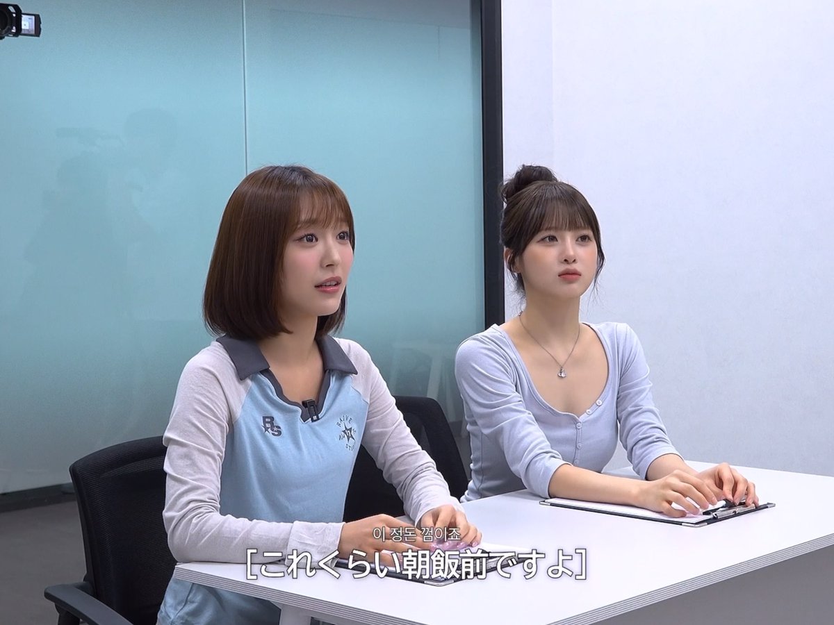 Two young East Asian women sit at a white table in a studio setting; the woman on the left wears a blue and white polo shirt and speaks, while Japanese subtitles read '이 정도 길이죠 [これぐらい朝飯前ですよ]'.