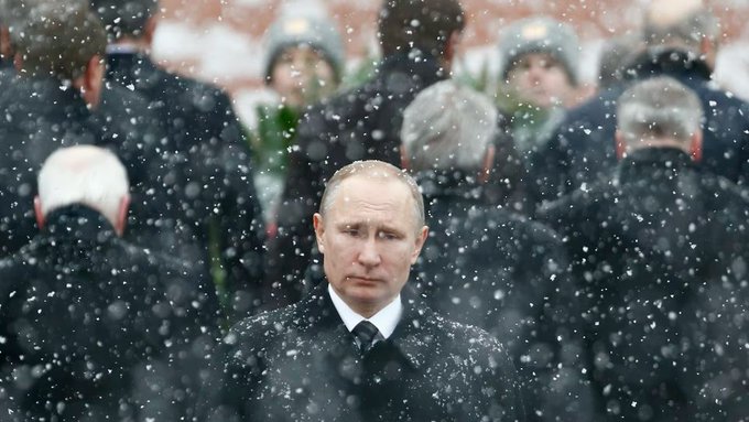 A stern-faced Vladimir Putin stands solemnly in falling snow, flanked by security personnel in dark coats, evoking the harsh winter conditions referenced in the post about Russia's energy attacks on Ukraine's power grid as temperatures plummet.
