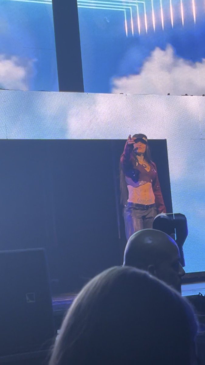 A female performer wearing a blinder and a long jacket sings on a large stage with a blue backdrop, viewed from the audience.