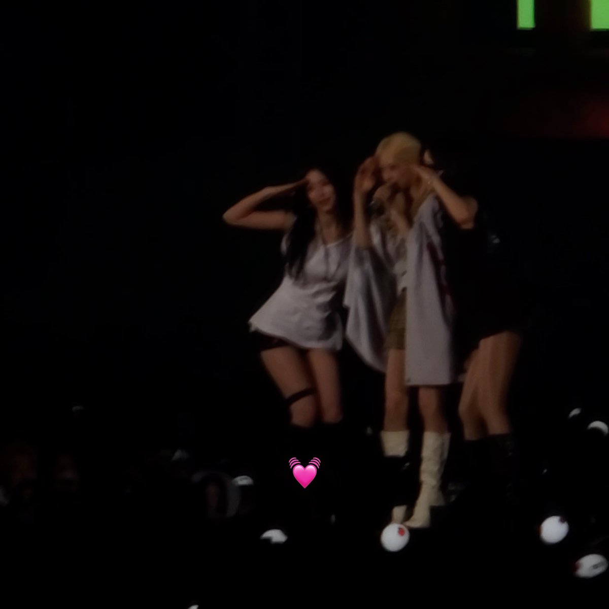 A blurry, dark photograph of three women in light-colored outfits performing on a stage, posing with their hands near their faces while holding a microphone, with a pink heart-emoji animation overlaid.