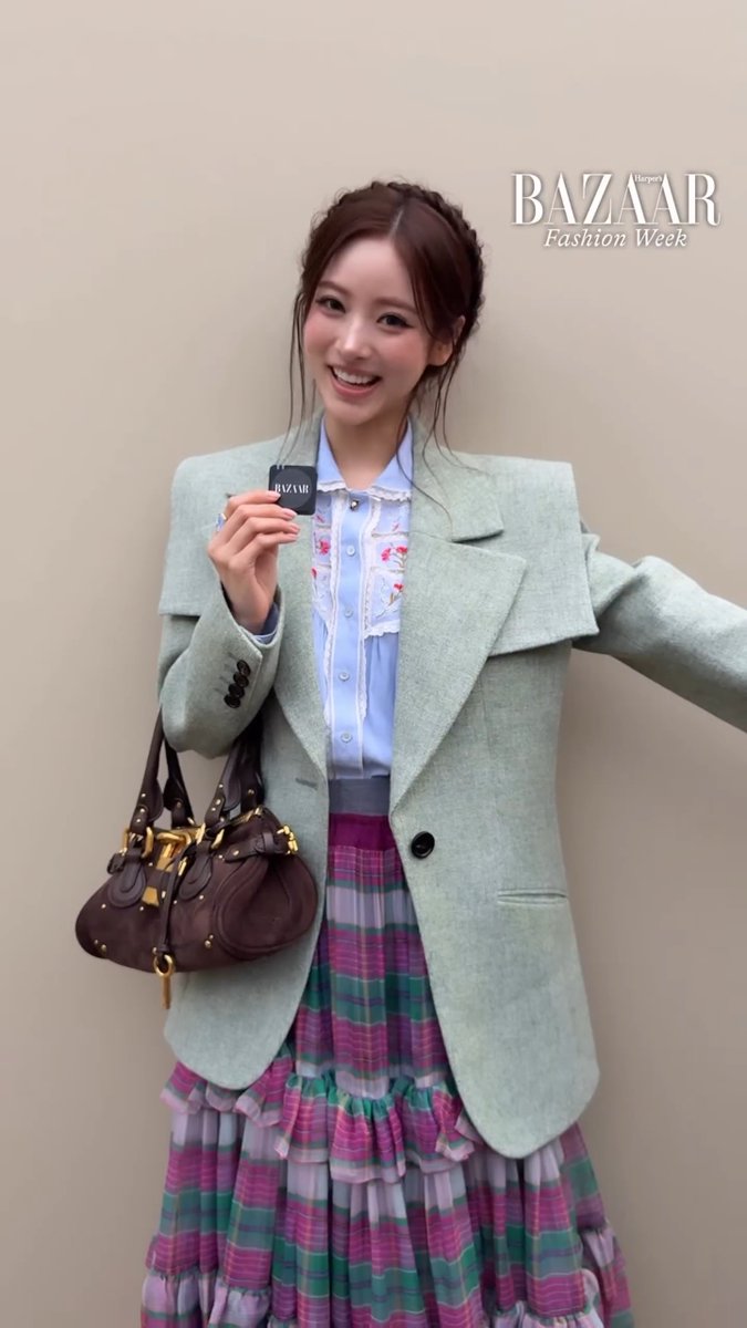 Editorial fashion photo with a 'BAZAAR Fashion Week' watermark showing a smiling woman wearing a light green oversized blazer over a blue floral blouse and a tiered purple and green plaid skirt, carrying a dark brown Balenciaga-style bag.
