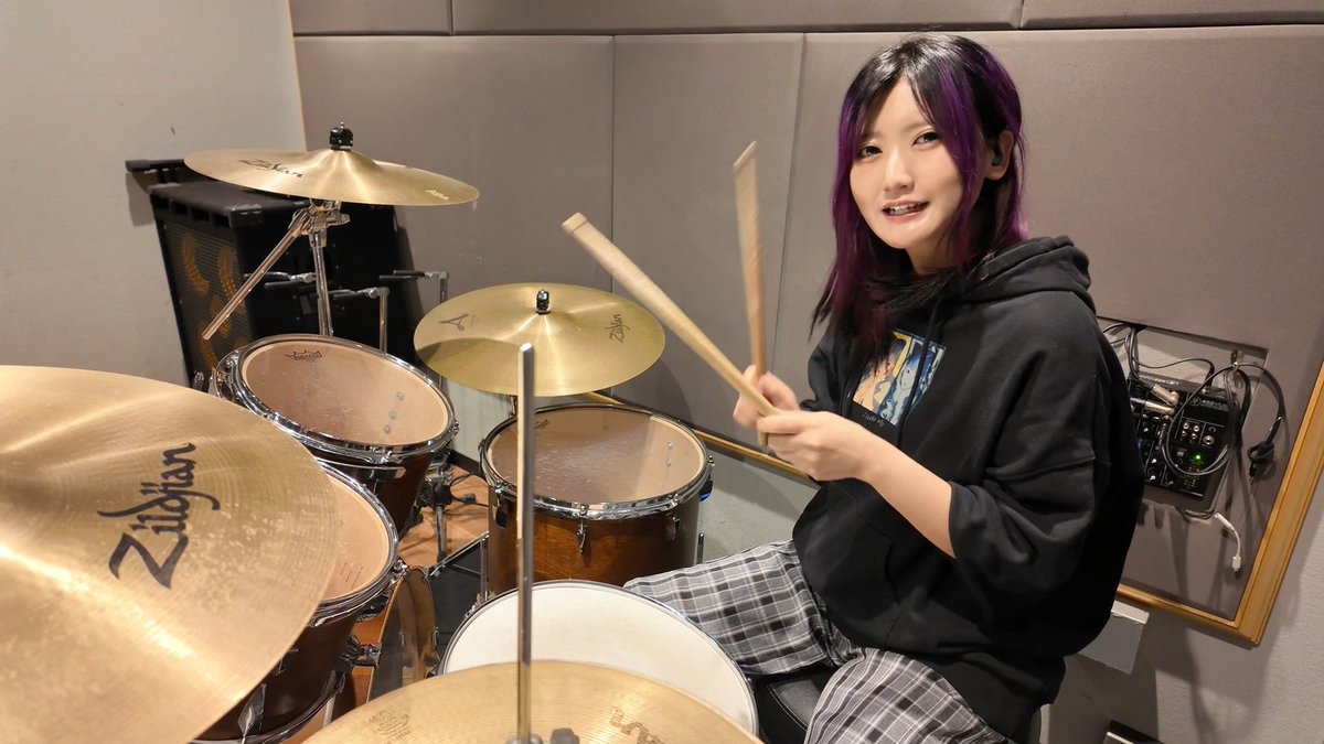 A drummer in her light purple hair afro-style is on a drum kit with Zildjian cymbals.

The image shows a young woman with purple-tipped hair sitting behind a drum kit featuring multiple Zildjian cymbals, holding drumsticks while looking at the camera in what appears to be a soundproof practice room with audio equipment in the background. This personal photo seems to showcase her musical background, contrasting with the announcement about her new venture into pachinko/pachislot entertainment work at pachinko parlors.